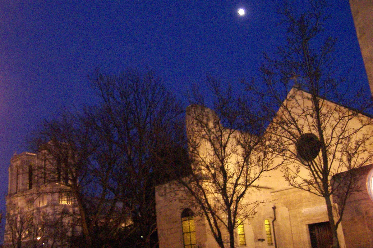Saint-Julien-le-Pauvre lit with warm light under a deep blue evening sky, with the silhouette of Notre-Dame behind and a moon above.